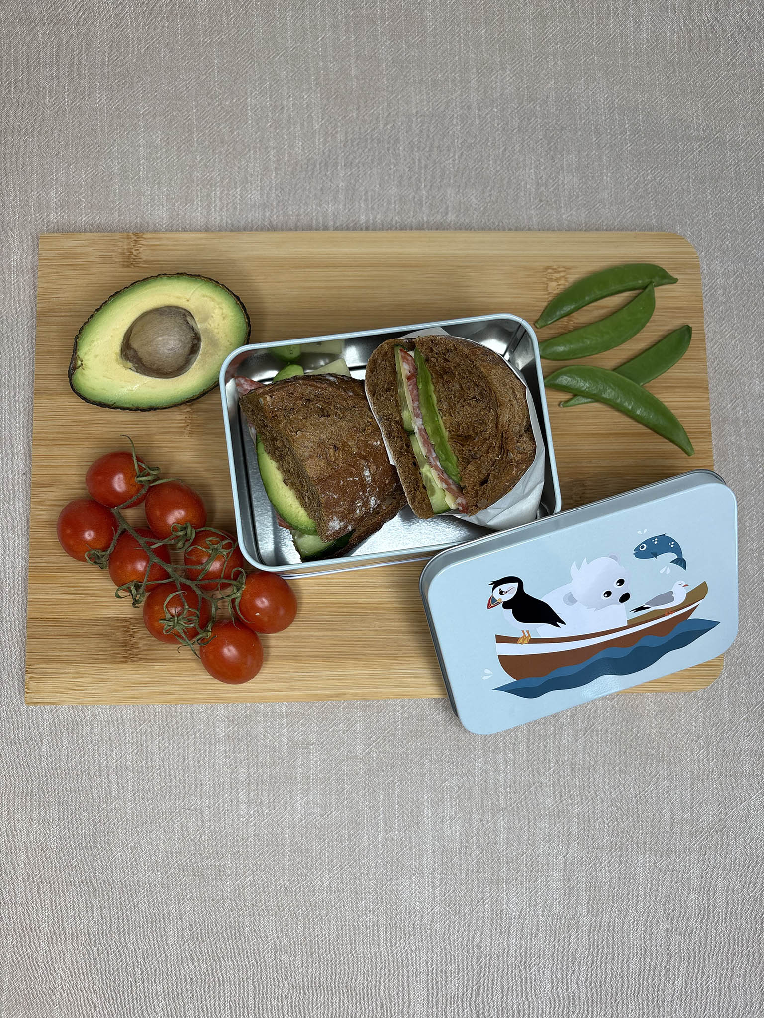 Blafre matboks i metall med lundefugl- og isbjørnmotiv, på trebrett med avocado og tomater.