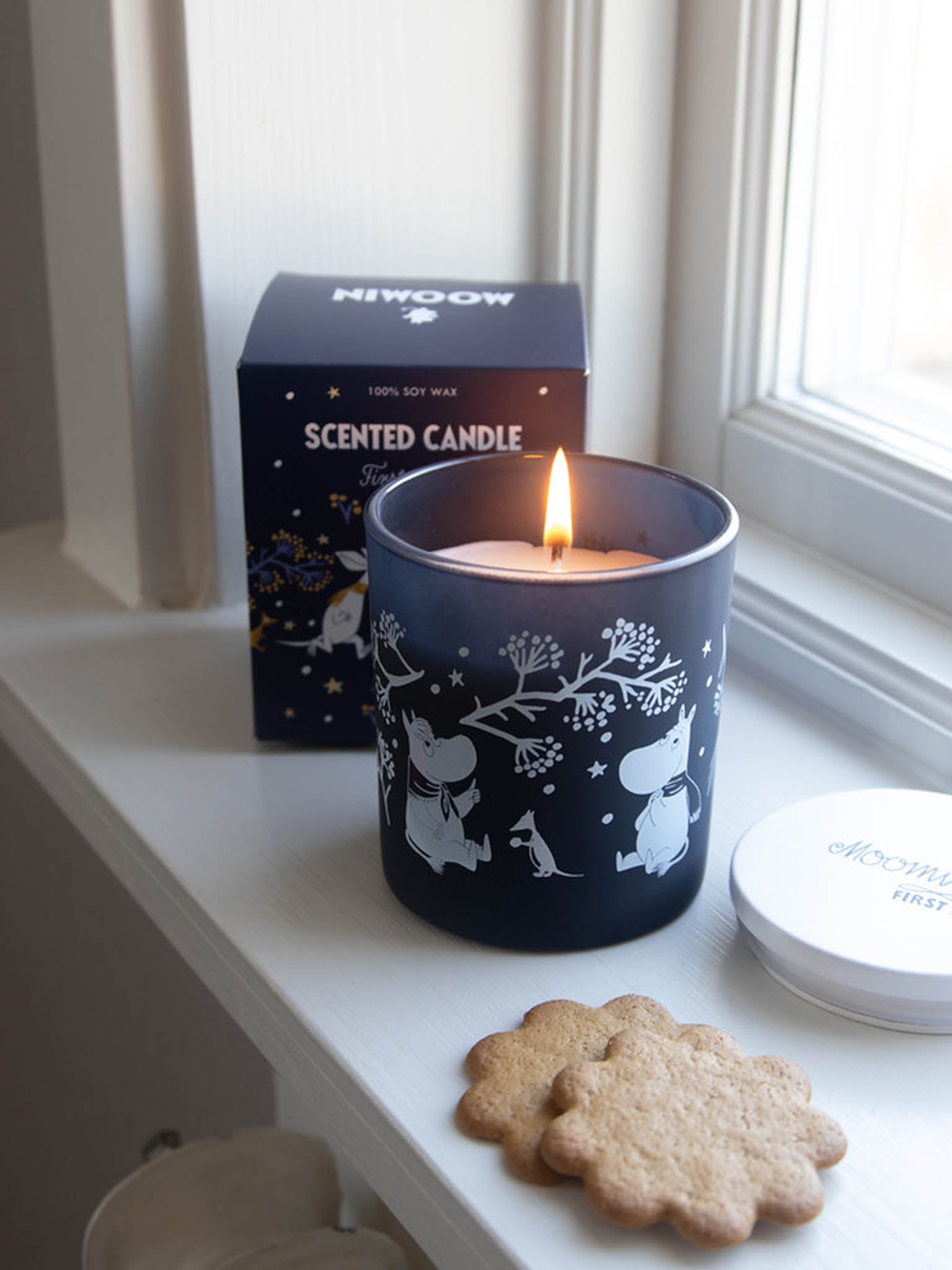 Tændt duftlys MOOMIN First Snow i blå sylinder med hvite figurer, ved siden af eske
