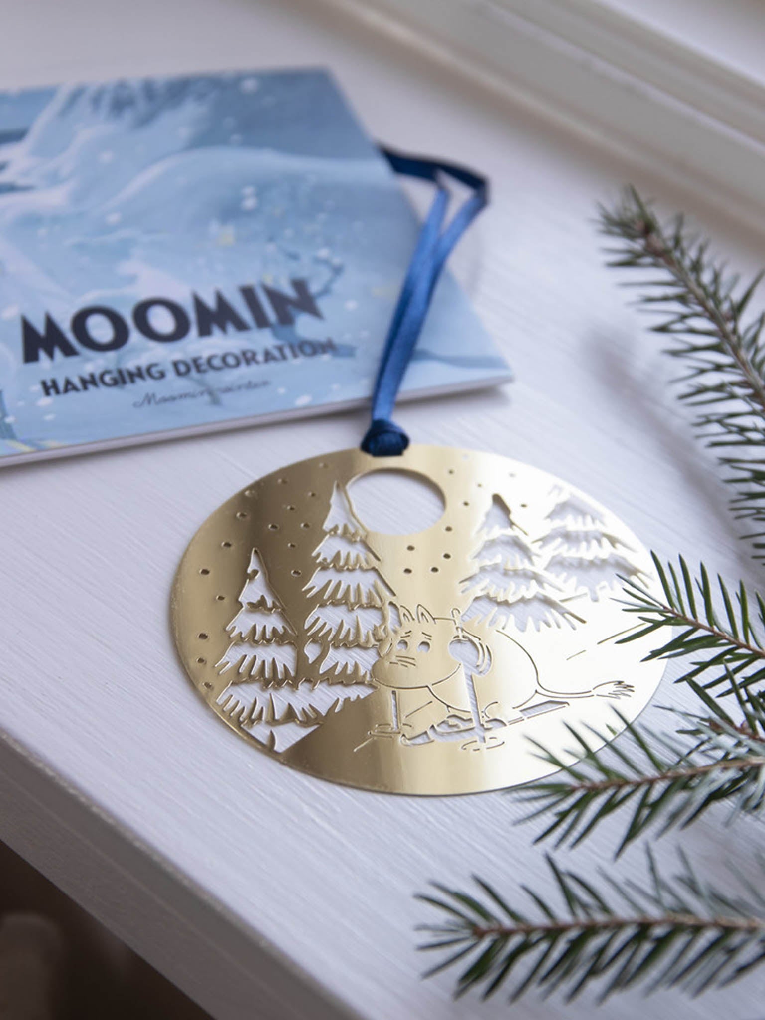 Pyöreä Moomin-ripustuskoriste kultasävynä, valkoinen puukuva ja Moomin, sininen nauha